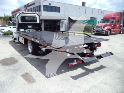 Flatbed wrecker tow truck on a rollback carrier with rear ramps and lights visible; Peterbilt dealership in background
