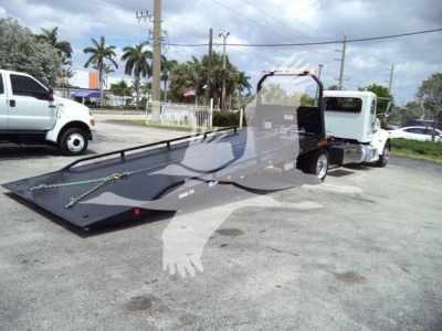 2018 Peterbilt 337 rollback tow truck with flatbed in a parking lot, side view showing the long tilt bed and white cab.