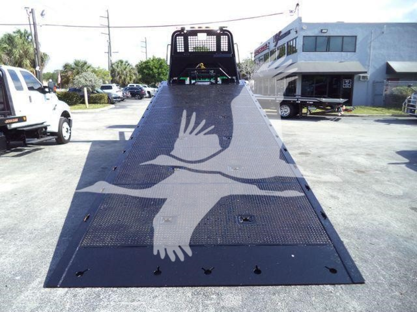 Truck flatbed ramp with a white bird emblem; rear view shows loading deck and black mesh, Freightliner M2 106 rollback vehicle in background.