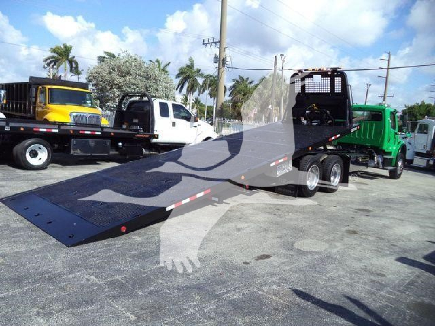 Flatbed tow truck with a long ramp in foreground and other trucks in background; Freightliner M2 106 trailer/rollback setup visible
