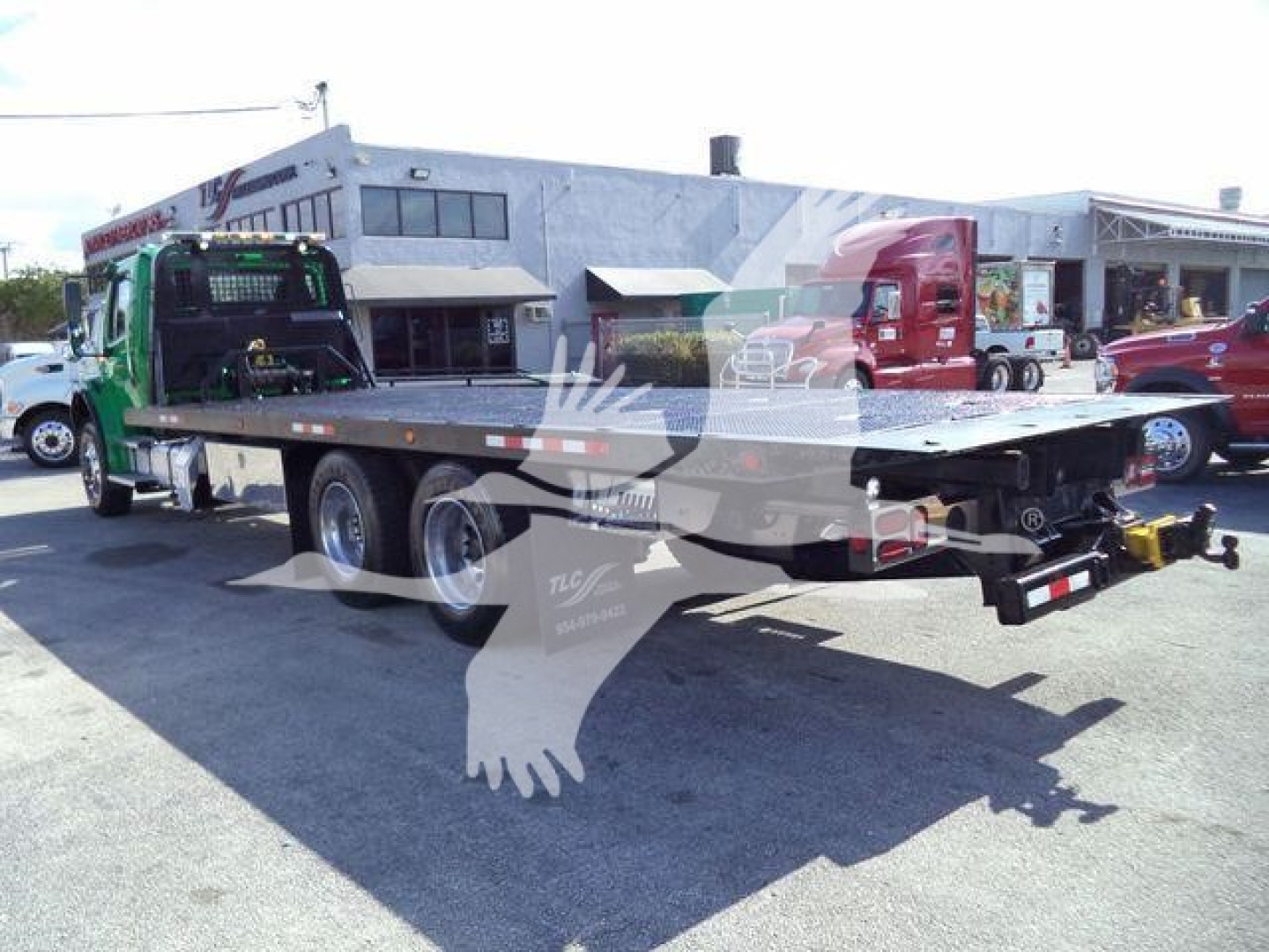 Flatbed tow truck attached to Freightliner M2 106 with green cab and extended bed, rear view showing hitch and license plate area.