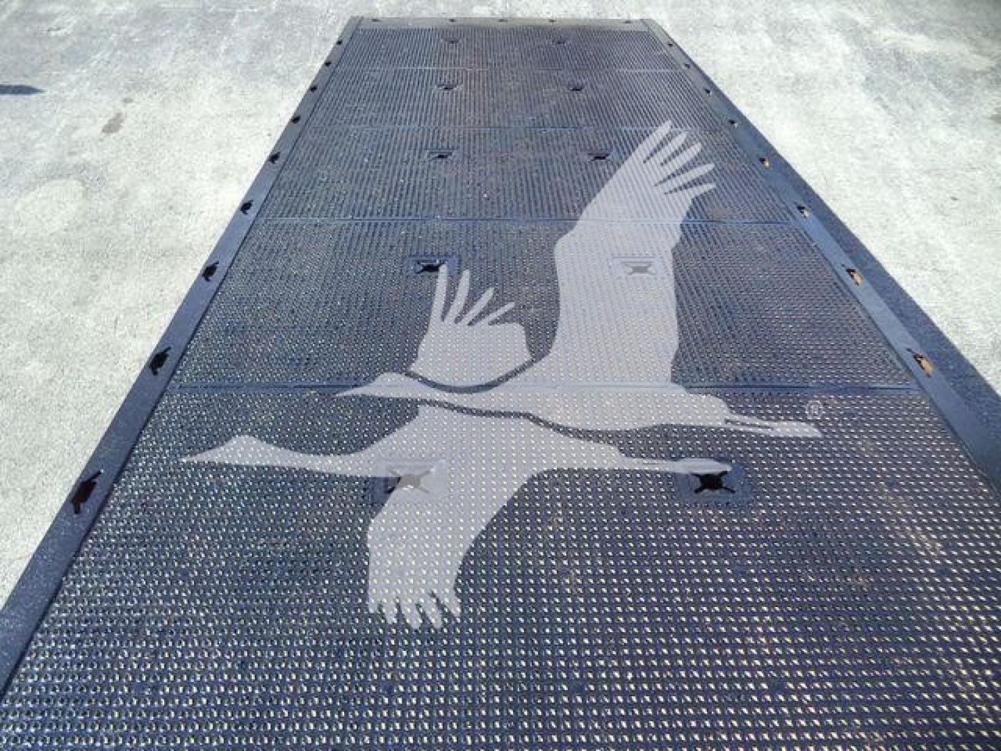 Flatbed trailer deck with a white bird graphic and textured metal surface, edge holes visible along sides.