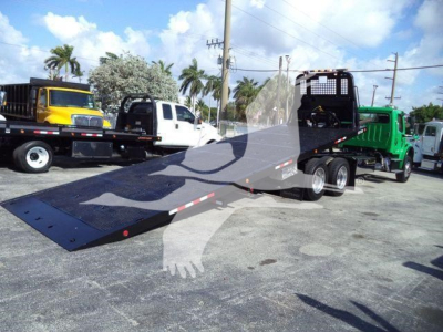 Flatbed tow truck with a long ramp in foreground and other trucks in background; Freightliner M2 106 trailer/rollback setup visible