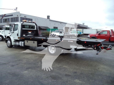 Flatbed tow truck with rollback equipment on a Freightliner M2 chassis, front cab visible, rear flatbed with wheel chocks, dealership lot in background.