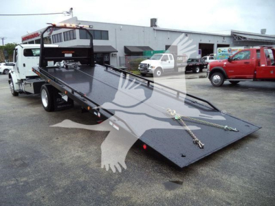 Truck bed/flatbed tow truck with a long black steel ramp in a dealership lot. Visible are two white and red pickup trucks, a building in background, and metal chains on the ramp surface.