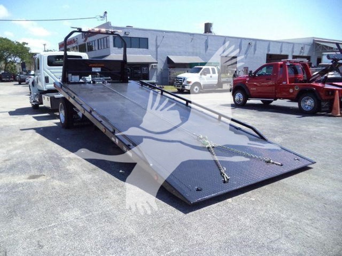 Tow truck rollback flatbed with ramp extended, black metal surface, hydraulic mechanism, truck cab in background.