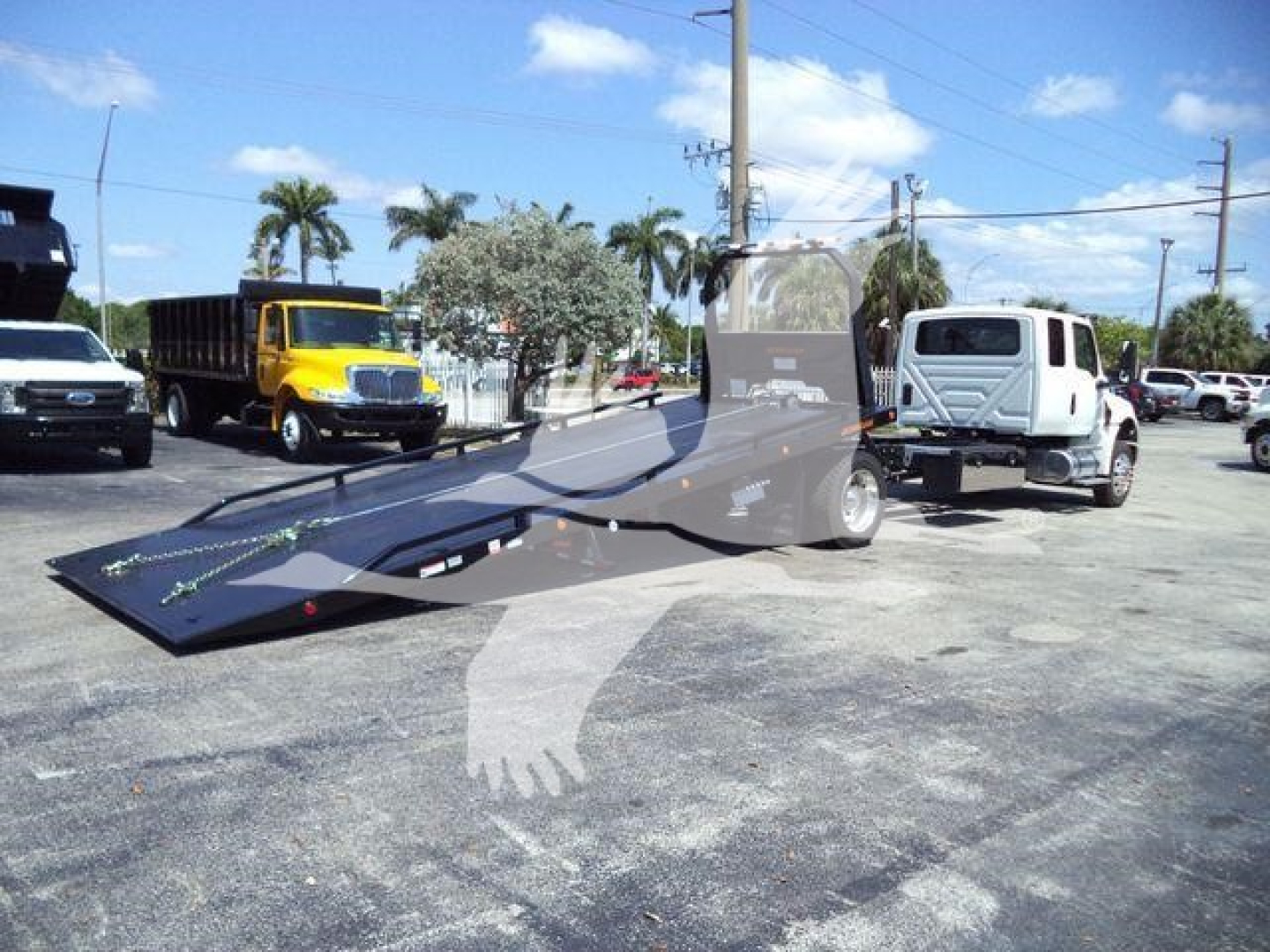 Truck with a flatbed rollback in a parking lot; yellow dump truck and white cab in background.