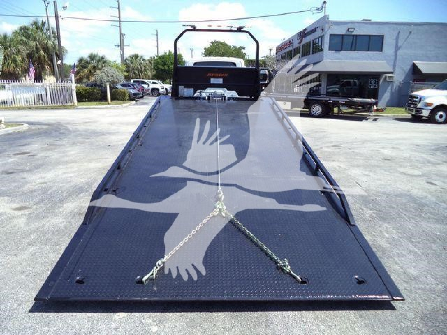 Flatbed tow truck trailer with a black diamond-plate surface; chains and hooks laid across; blue sky, surrounding buildings, and a carrier truck in background.