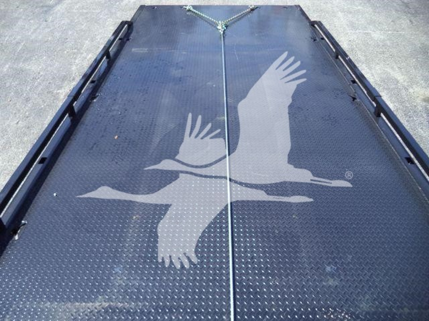 Flatbed trailer deck with textured steel surface and a white graphic of two soaring birds; there are tie-down rails along the edges and a center seam.