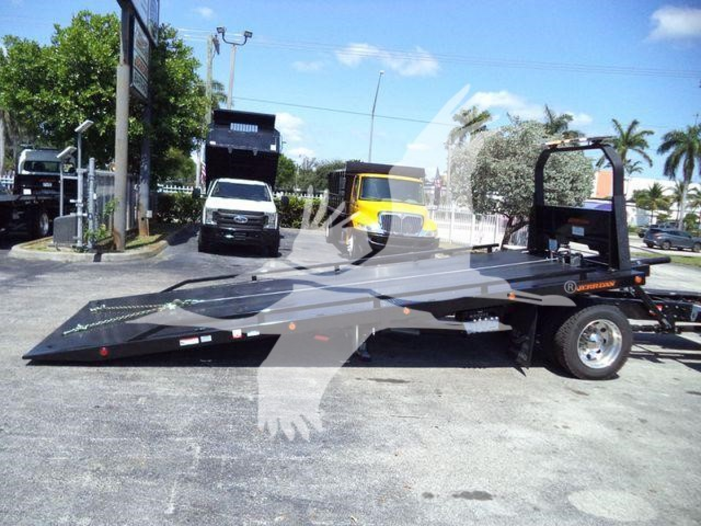 Flatbed rollback truck shown in side view with loading ramp, branding, and warning decals; background includes a white van, a yellow truck, and palm trees.