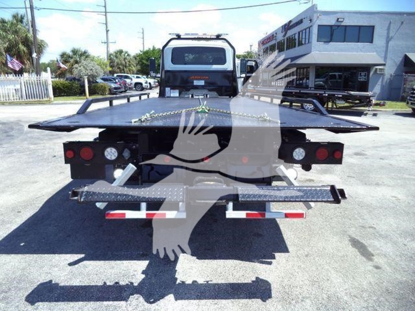 MV-style rollback truck flatbed showing rear view with hydraulic bed and taillights, white and black with red reflectors, dealership lot background.