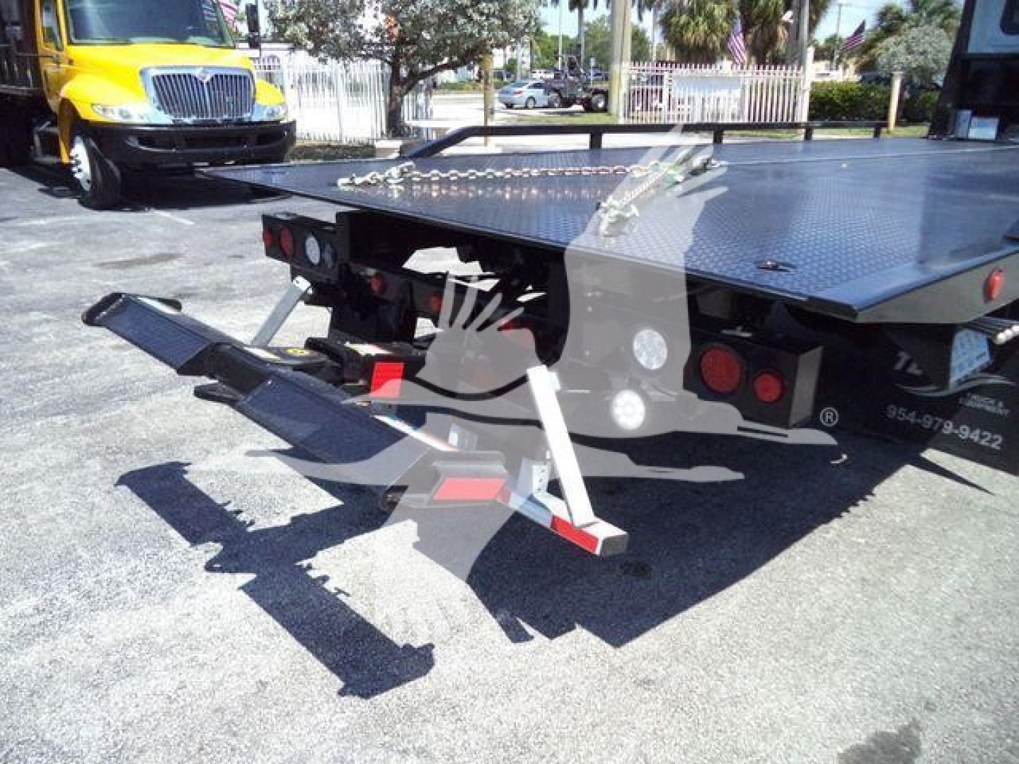 Trailer with hydraulic rollback ramp in lowered position, platform with diamond plate, rear view showing ramps and support legs, International MV truck in background.