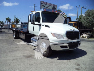 Truck bay showing a white International MV rollback truck with a long flatbed, front grille, and large side mirrors in a dealership lot.
