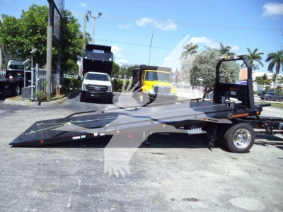 Flatbed rollback truck shown in side view with loading ramp, branding, and warning decals; background includes a white van, a yellow truck, and palm trees.