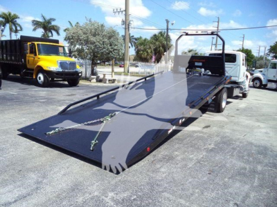 Truck with a flatbed ramp in a dealership yard, International MV rollback truck in background