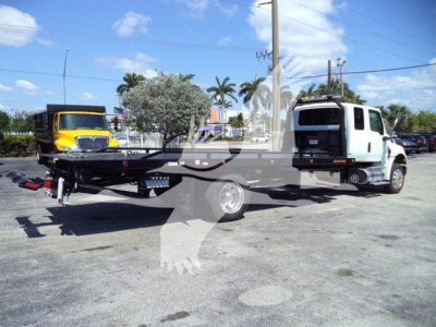 Truck with a rollback tow bed attached to a white cab; equipment on the platform with a yellow truck in background.