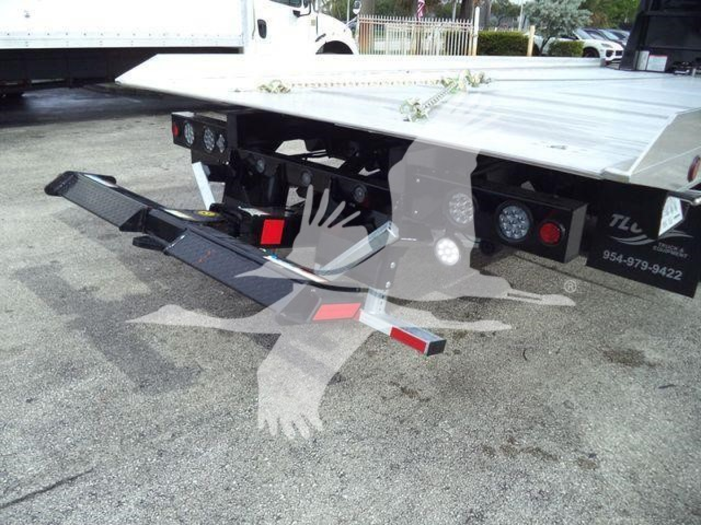 Rear/side view of a flatbed rollback truck with a black steel loading ramp and taillights; visible stickers show serial/phone numbers and a reflective logo.