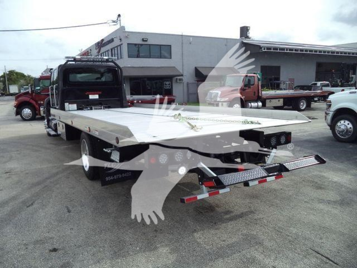 Flatbed tow truck rear view showing long aluminum deck and rear ramp with taillights and bumper, parking lot scene with other trucks.