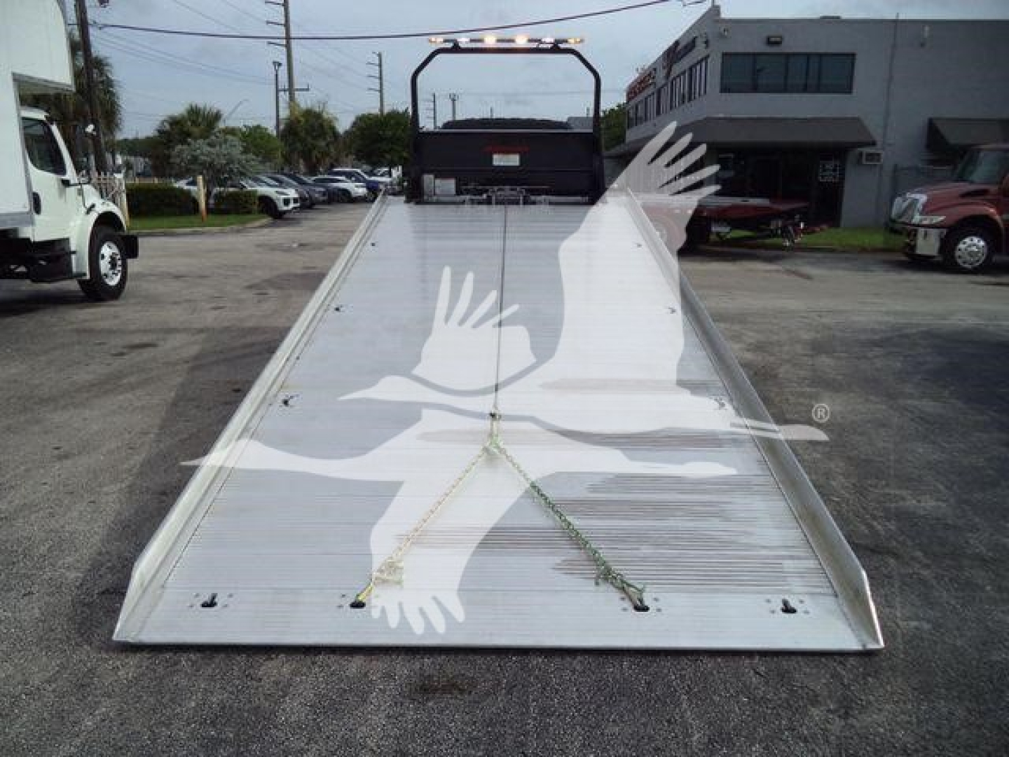 Flatbed tow truck bed with ramp and chain tie-downs; International MV flatbed in foreground, dealership lot in background.