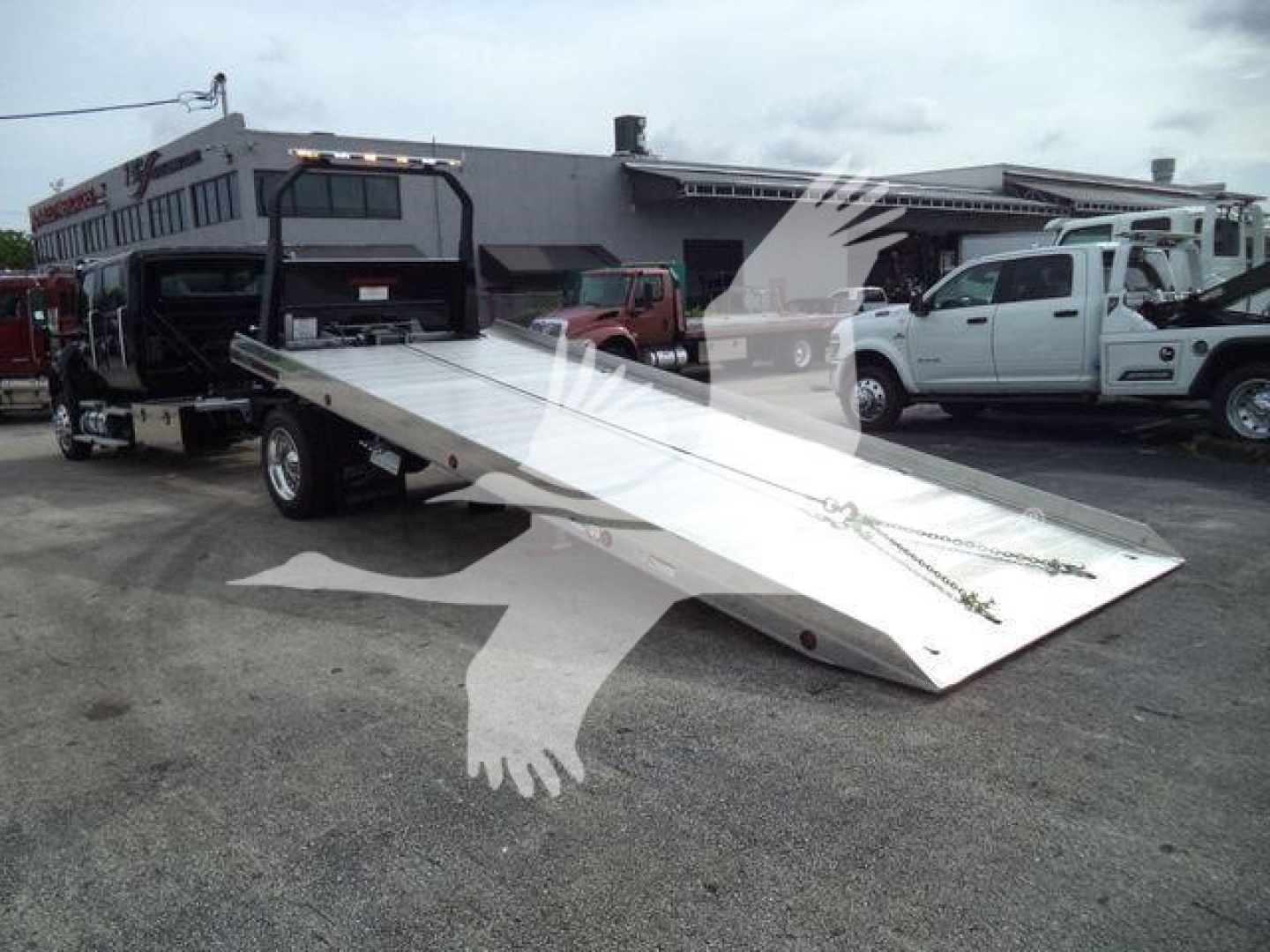 Truck with a silver flatbed ramp in a dealership lot; in background are other trucks and a building.