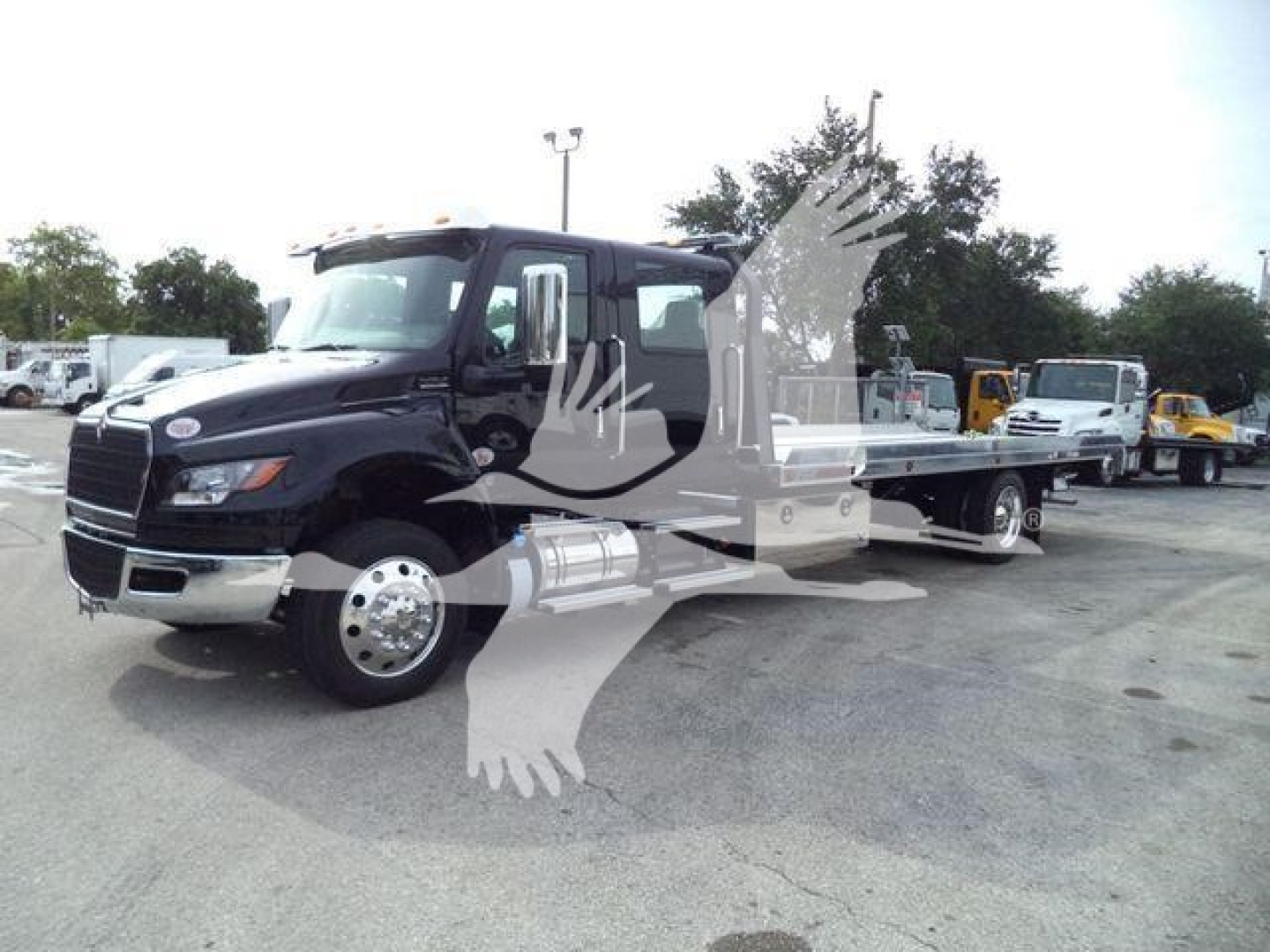Flatbed tow truck with dark blue cab, long chassis, front grille and chrome bumper, multiple wheels visible; image shows a rollback truck in a lot with other equipment in the background.