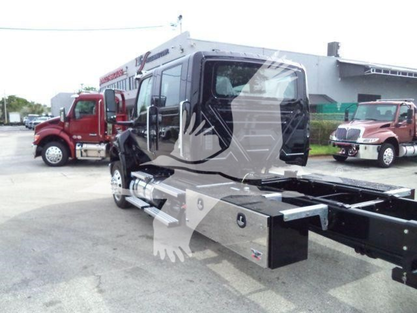 MV bedless view of a 2026 International MV rollback tow truck with a large side toolbox and chassis frame visible; other service trucks in background.