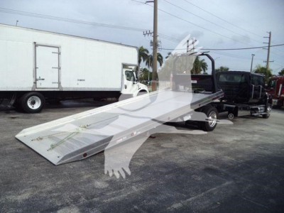 Truck with a long metal tow ramp or rollback bed laid out on the ground; background shows a white box truck and other vehicles.