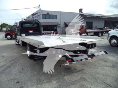 Flatbed tow truck rear view showing long aluminum deck and rear ramp with taillights and bumper, parking lot scene with other trucks.