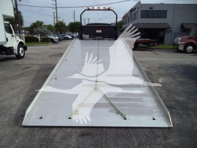 Flatbed tow truck bed with ramp and chain tie-downs; International MV flatbed in foreground, dealership lot in background.