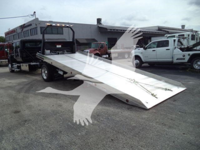 Truck with a silver flatbed ramp in a dealership lot; in background are other trucks and a building.