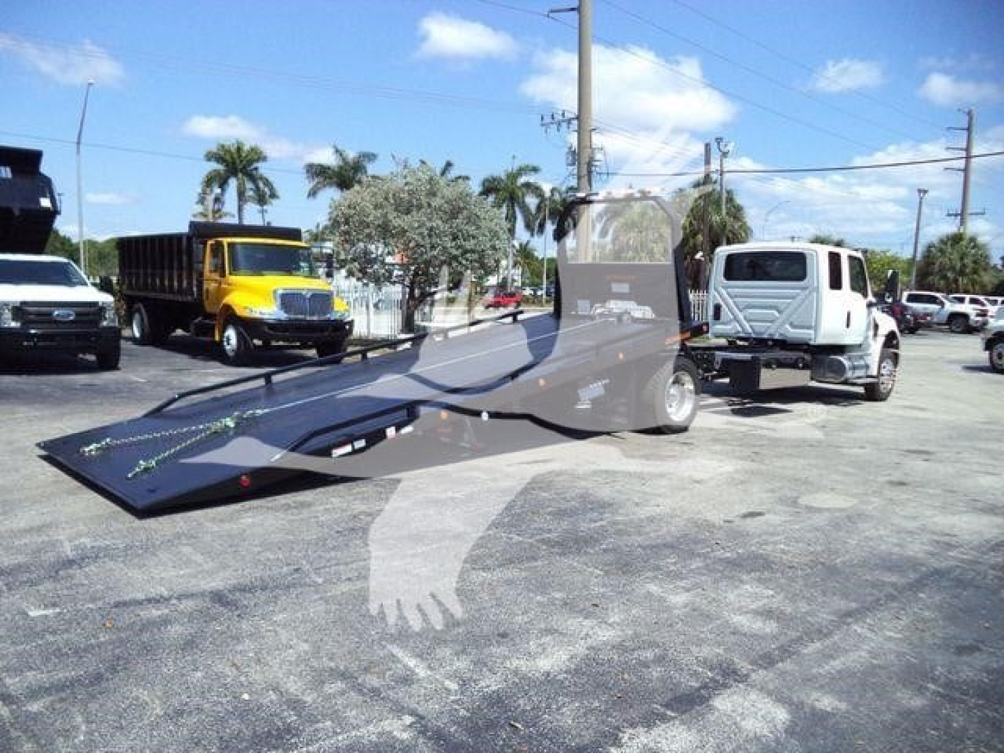 Truck rollback tow truck with flatbed in yard showing loading ramp and two white trucks in background.