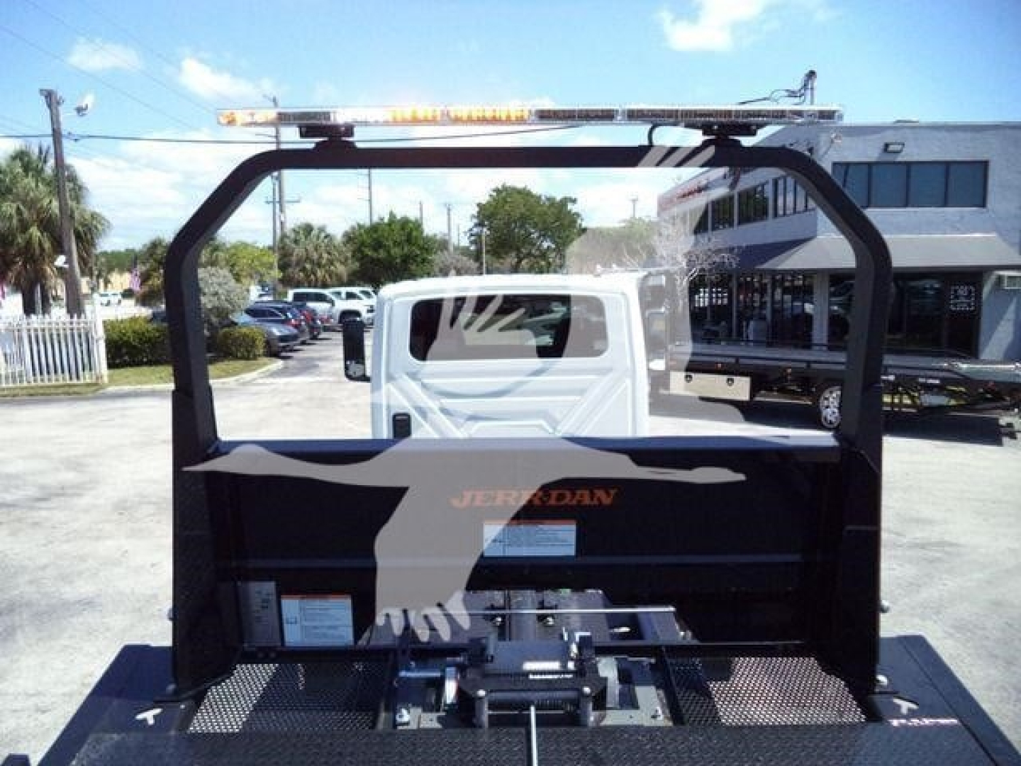 Truck bed with a Jerr-Dan rollback mechanism and warning light bar on top, seen from rear; visible white cab, black equipment frame, and pavement in background.