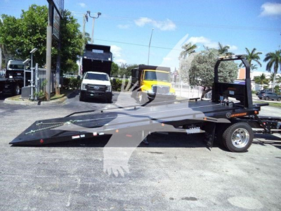 Flatbed rollback tow truck in a dealership lot showing a black aluminum deck with ramps, International MV truck line; visible text on a sticker near the rear wheel area and on the towing equipment.
