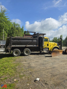 Side view of a yellow 2003 Freightliner FLD120 SD dump truck with a plow attachment. The truck has a black dump bed, three rear axles, and rugged tires. Open sky and trees in the background.
