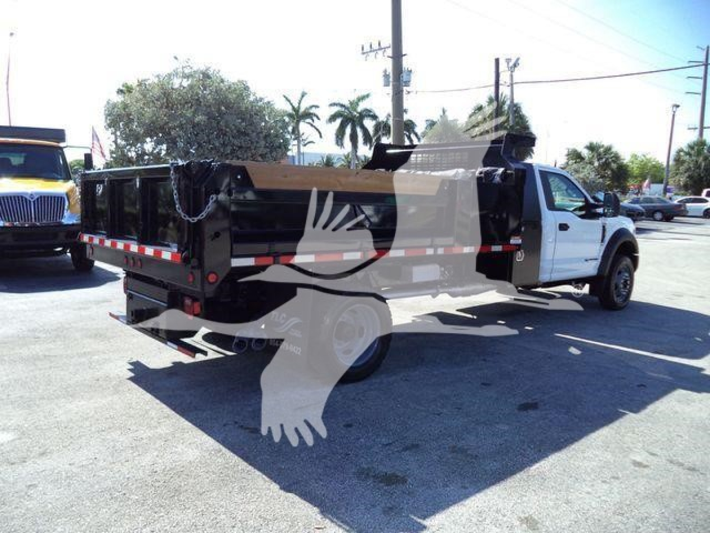 Dump truck bed with white Ford F-550 pickup truck in background, side view showing hydraulic dump bed and chain mechanism.
