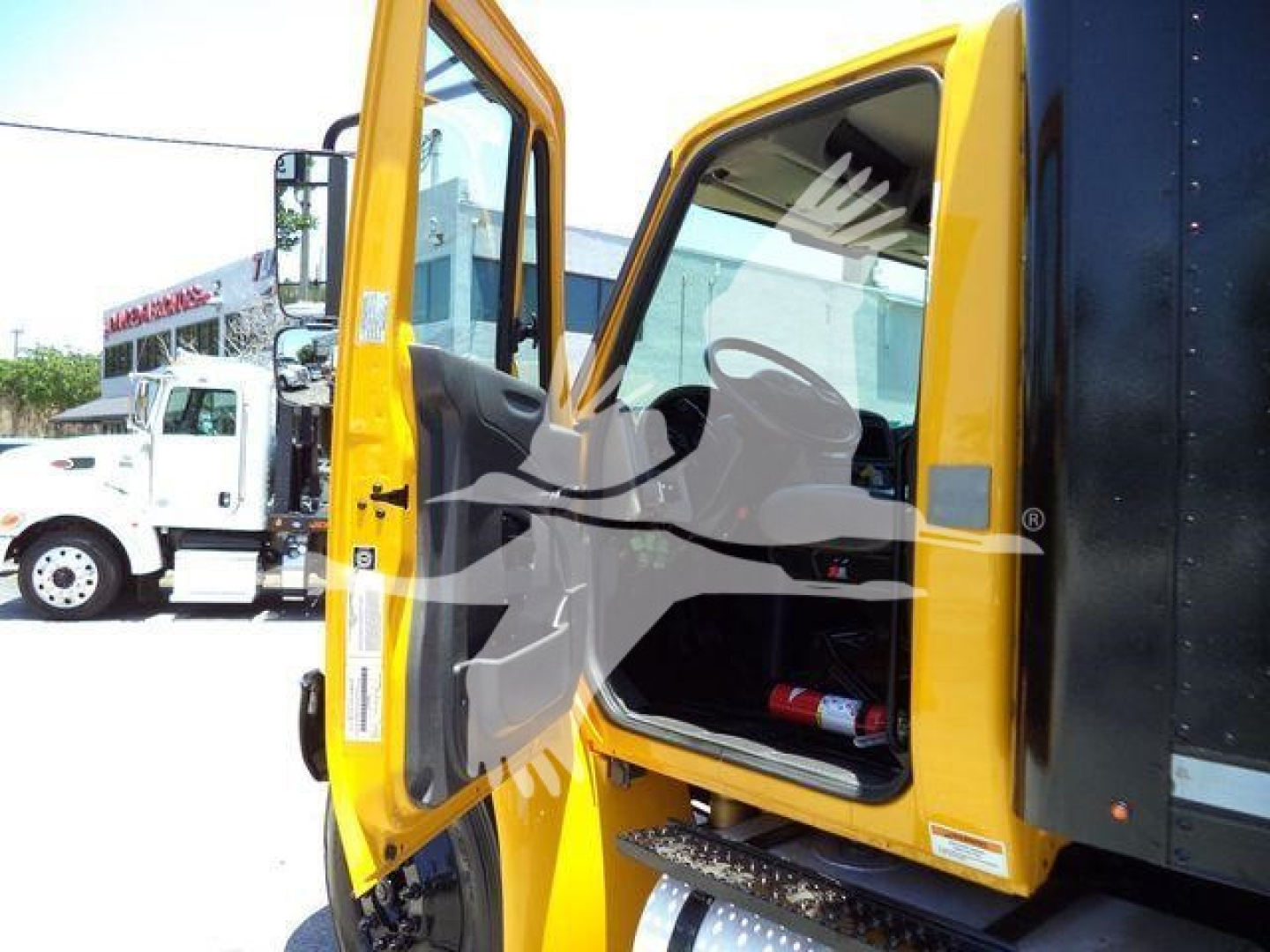 Truck cab of a 2019 International Durastar 4300 with the door open, showing the interior and step access, yellow cab color, and a nearby white truck in background.