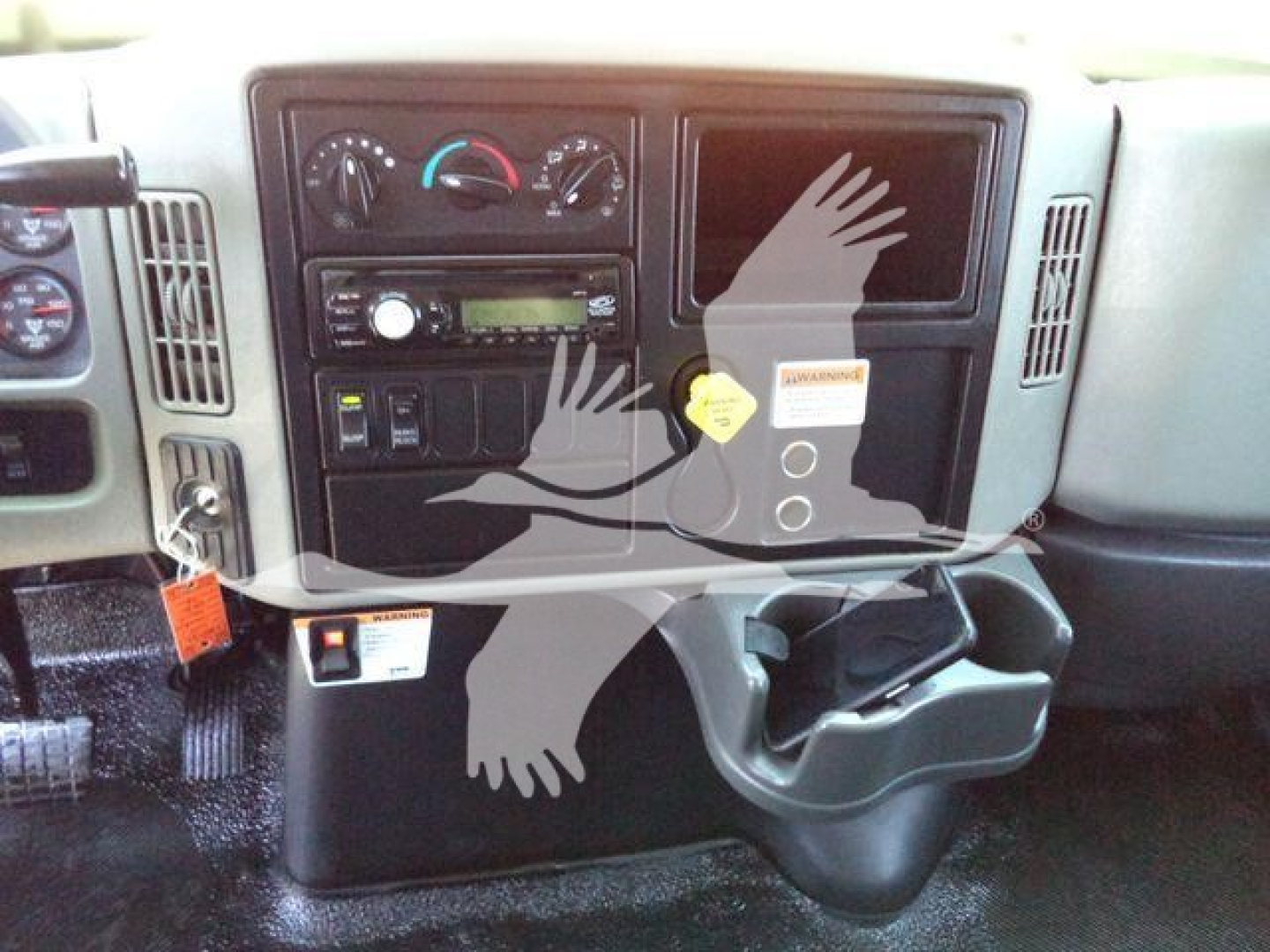 Truck dashboard view showing climate controls, radio, and empty display with cup holder; partial steering wheel and ignition to left; warning tag on center panel.