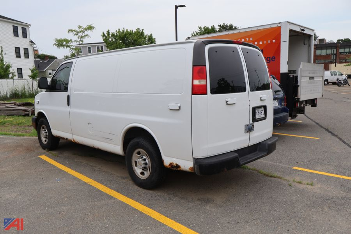 Rear and side view of a white 2005 Chevrolet Express cargo van with slight rust on wheel well, black bumper, passenger side sliding door, and orange vehicle in background.