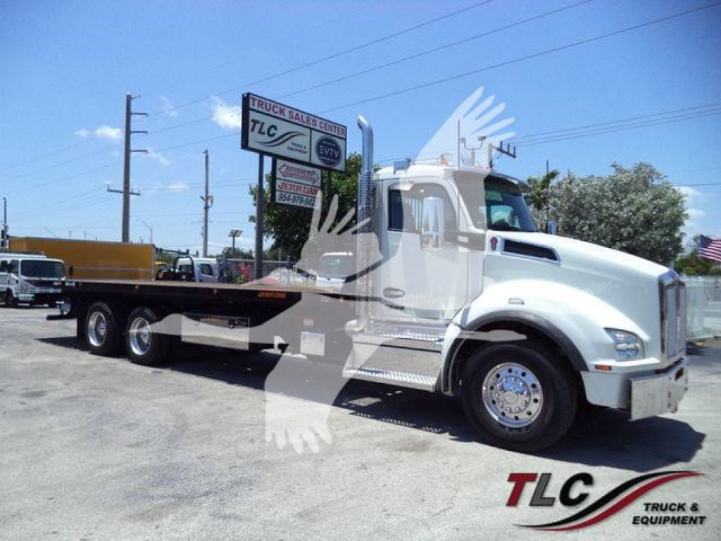 Kenworth T880 rollback truck in side view showing flatbed/rollback bed and chrome wheels with dealer signage in background.