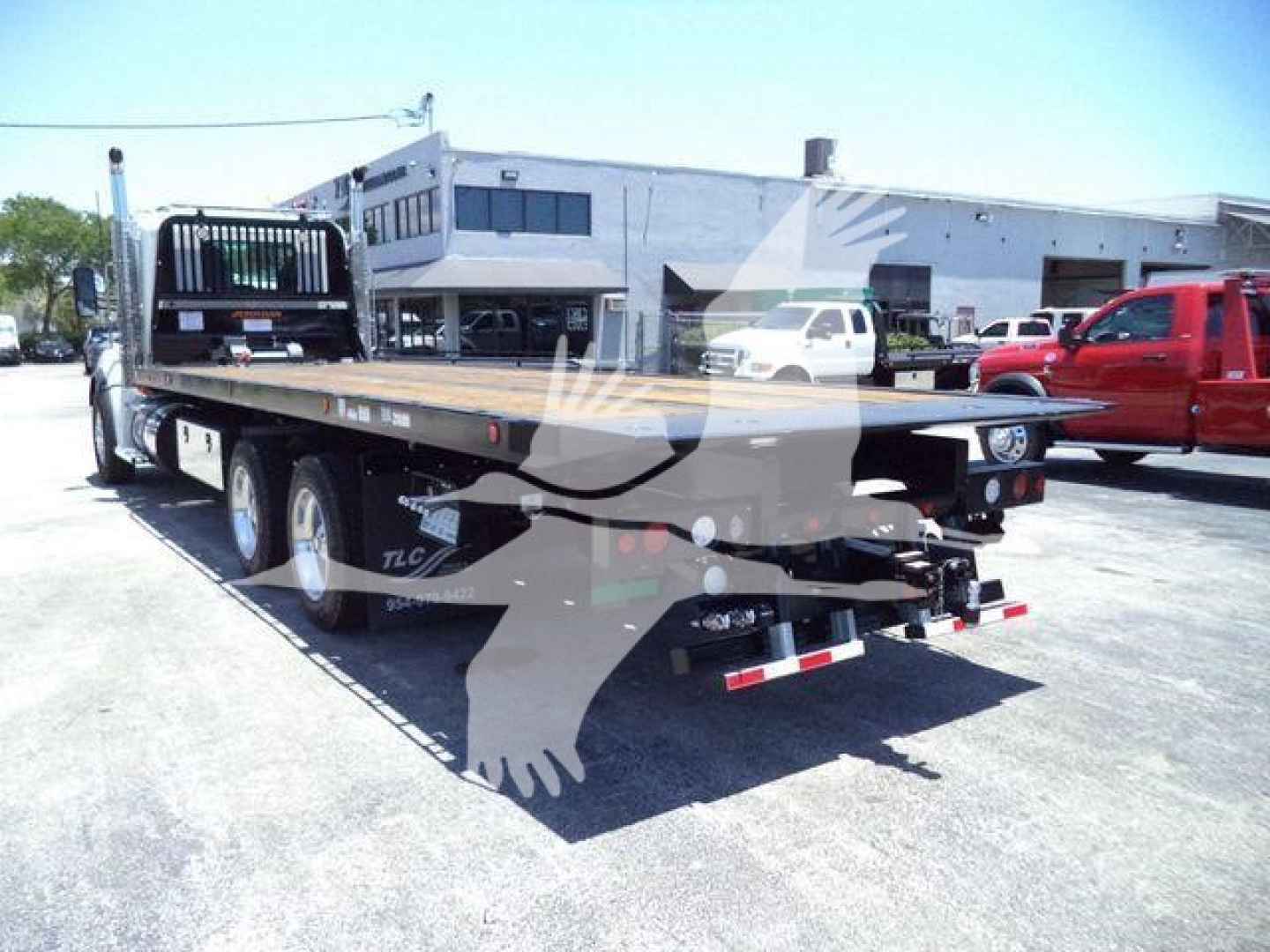 Kenworth T880 rollback truck in a lot showing flatbed with rear bumper and taillights; rear view with dual rear axles, equipment and dealership building in background