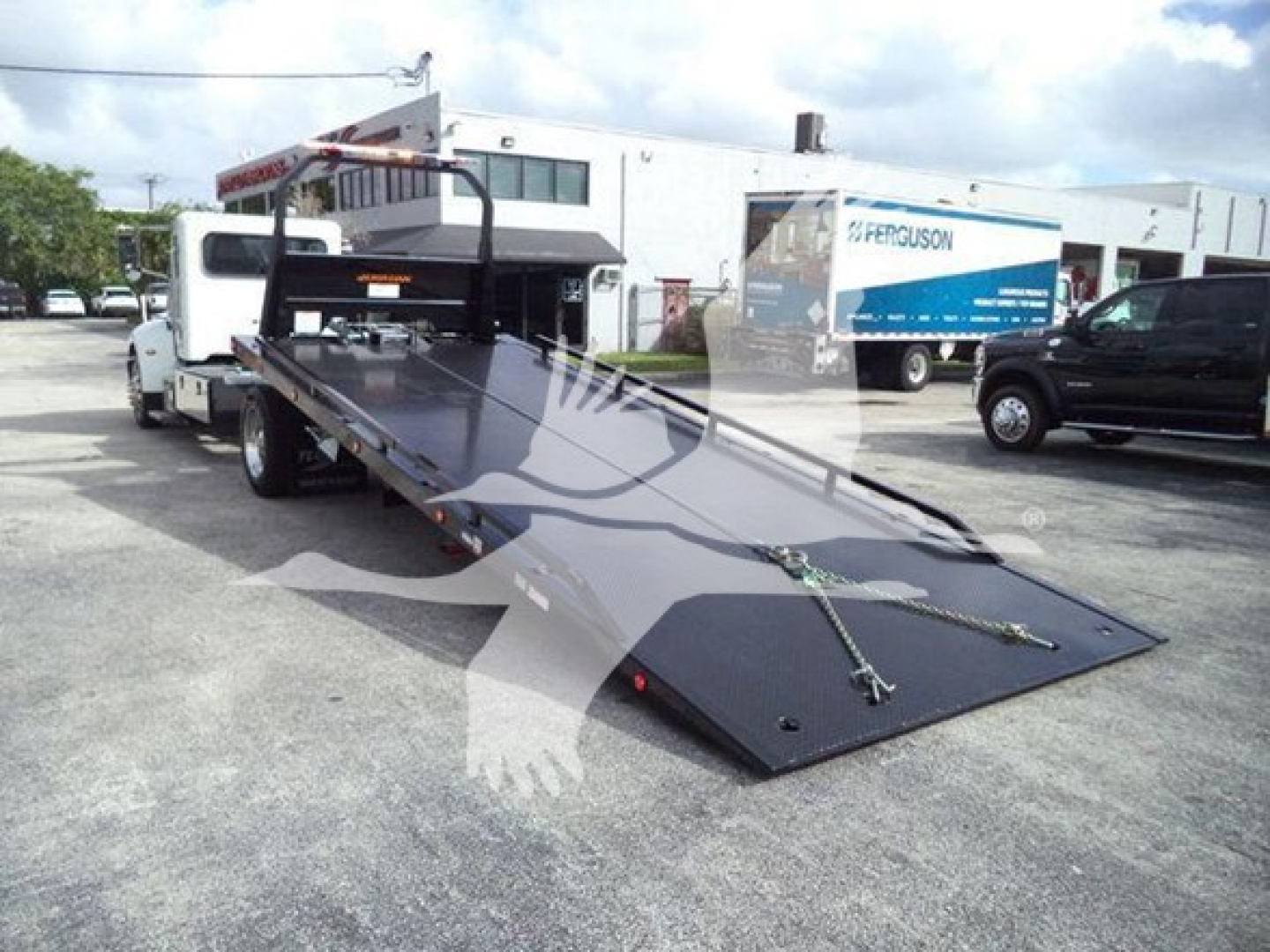 Truck bed with a long black + metallic rollback ramp attached to a white Peterbilt truck in a dealership lot.