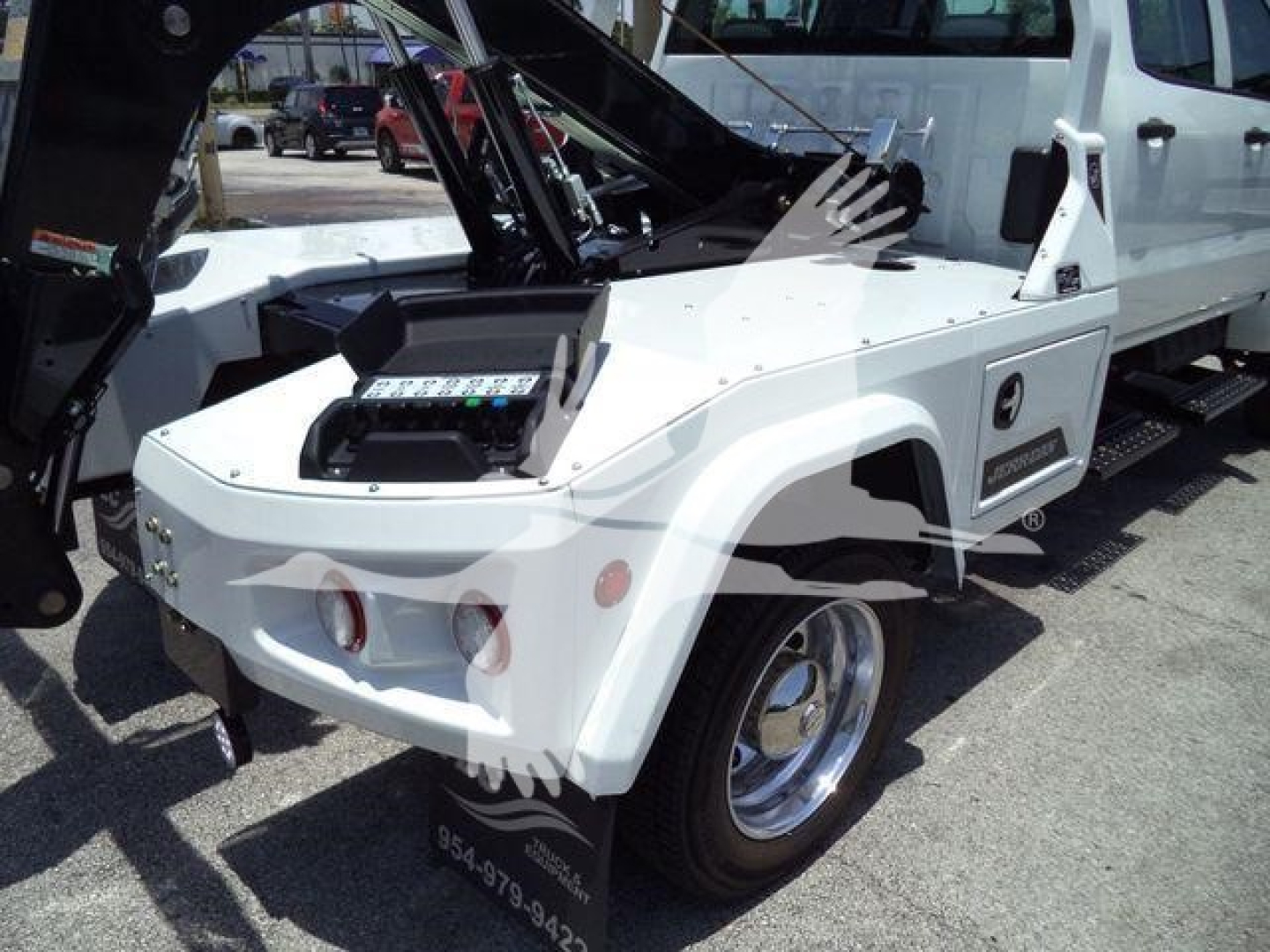 Truck with winch equipment visible on front fender and control panel in flatbed area; white vehicle with black hydraulic arms and chrome wheel hub.