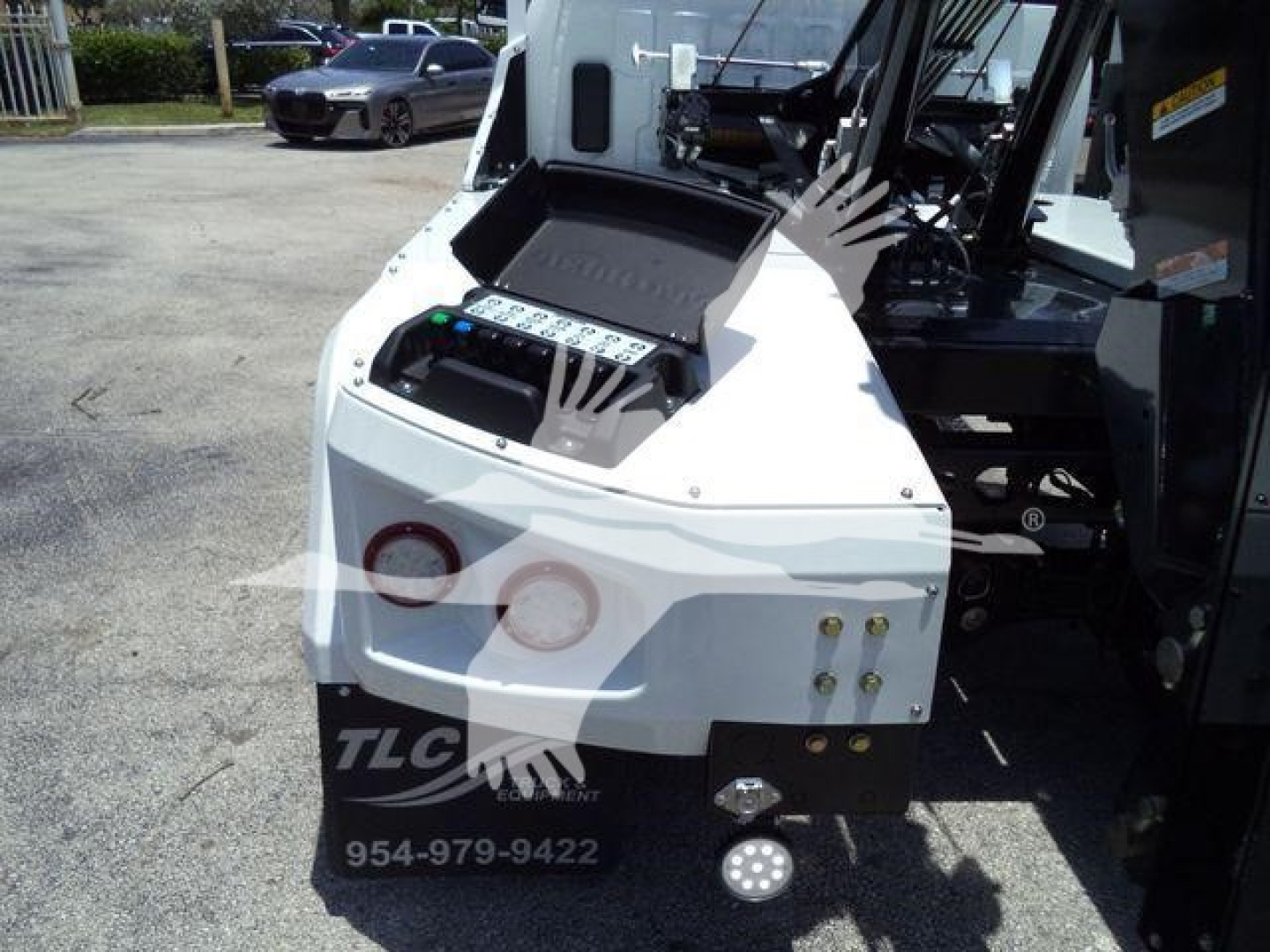 Truck winch control panel and front of a white International CV truck. Visible labels: stock number J19799FM, category Winch, type Trucks, new/used New. TLC contact number 954-979-9422 on the bumper area.