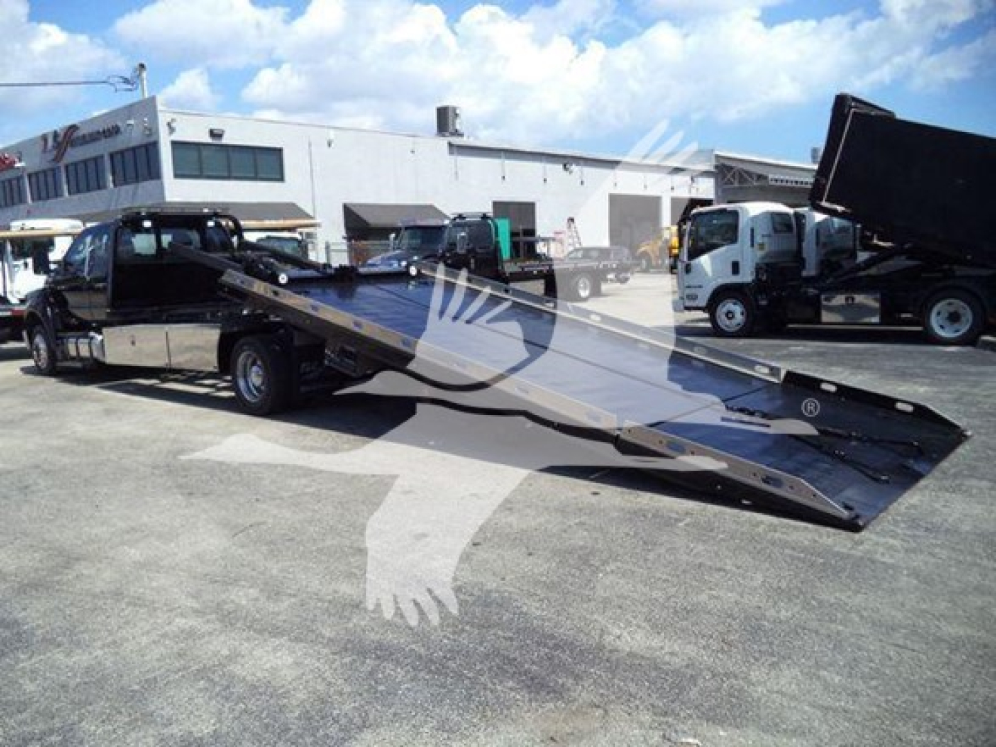 Ford F650 rollback tow truck with a long low-profile flatbed, seen in a lot with other trucks and a building in the background.