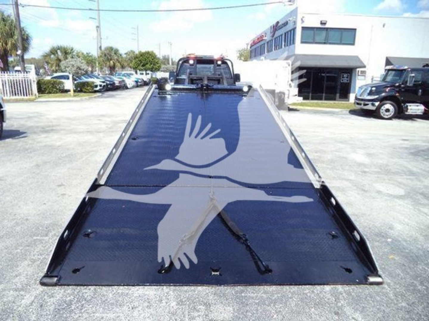 Flatbed tow truck with a black diamond-plate platform and white bird graphic. Front cab visible in distance under clear sky.