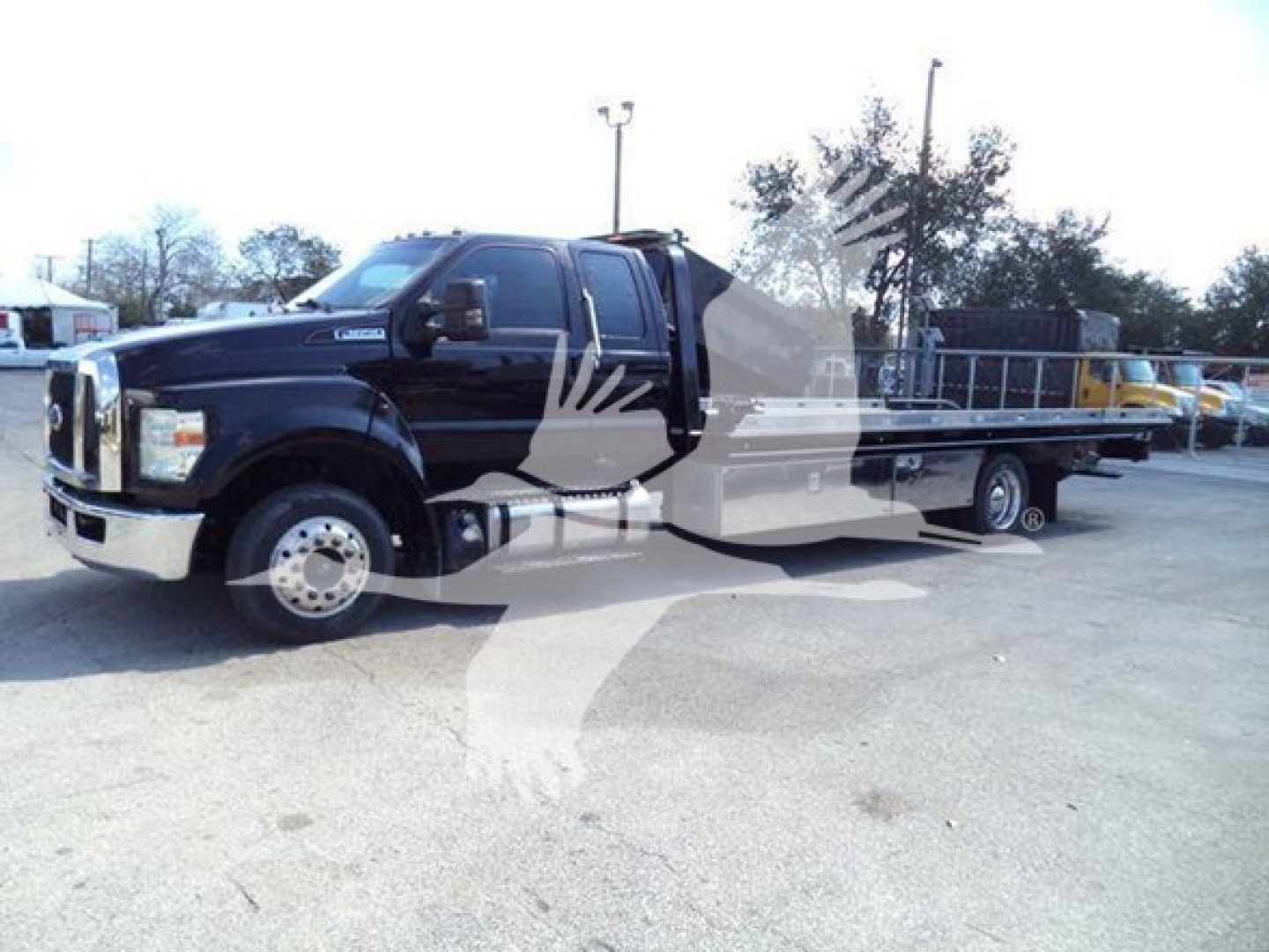 Ford F650 rollback truck in black with flatbed, side view showing extended cab, chrome front bumper, dual rear axles, and truck dealership lot in background.