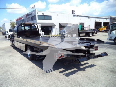 Truck rear view showing a flatbed rollback tow truck in a lot with a white building in the background.