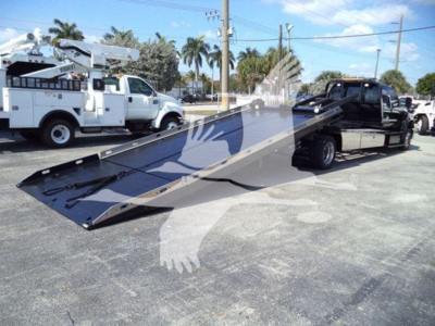 Ford F650 rollback truck with a tilted flatbed showing the loading ramp and tow truck in a parking lot.