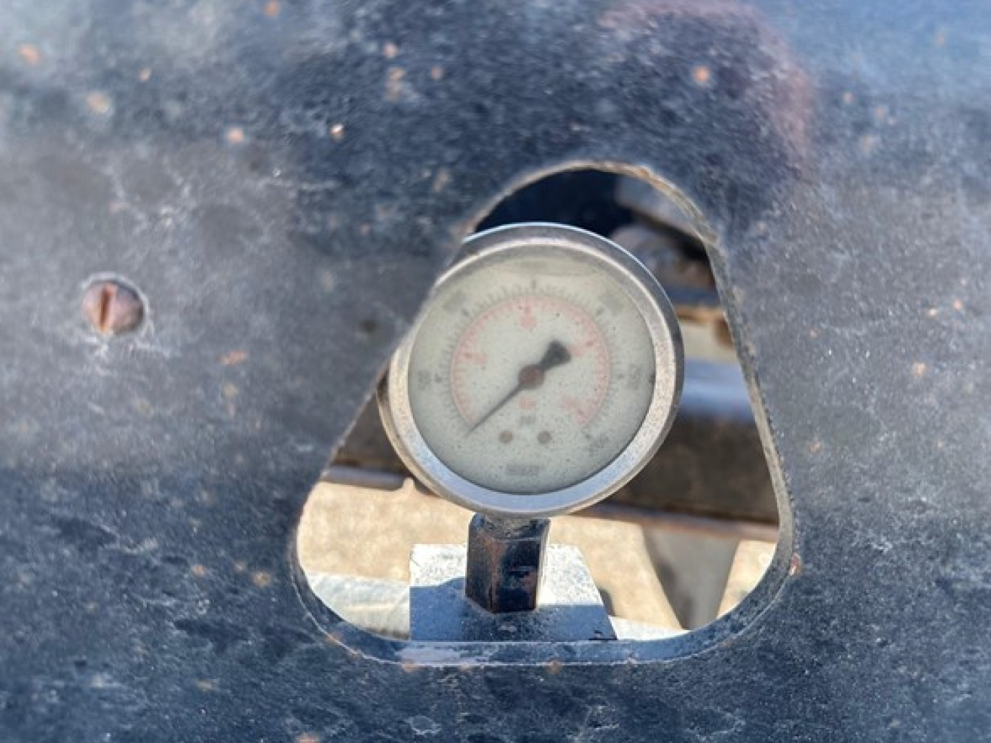Trailer gauge visible through a cutout in the side of a lowboy trailer showing a round dial gauge mounted on a metal bracket.