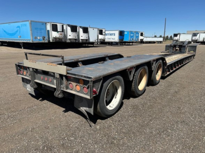 Lowboy trailer showing multiple axles and tires with a pony motor compartment at the front, bay area and backdrop of trucks in a yard.
